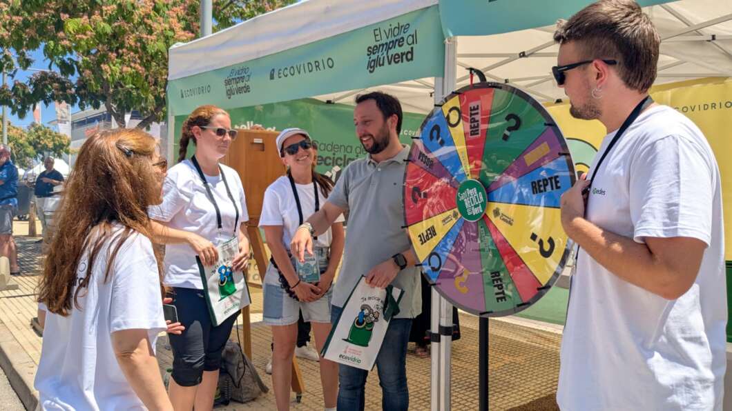 Group of people in white shirts gathered around a colorful prize wheel at an outdoor eco booth with banners in the background.