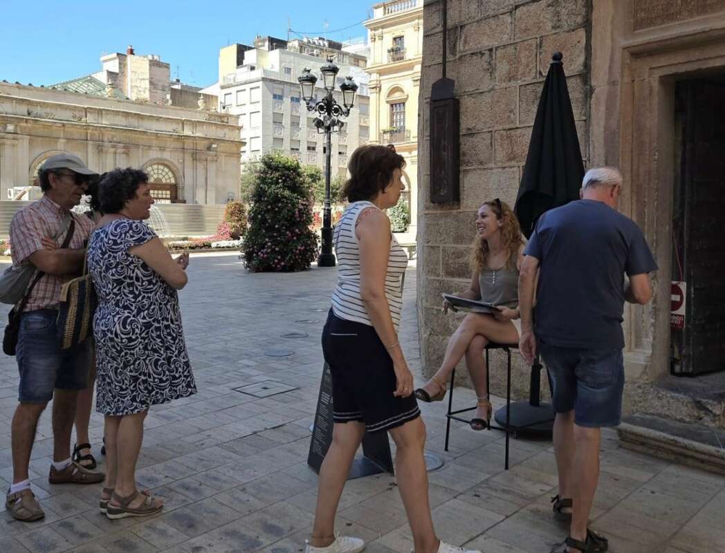 A tour group of five people listens to a guide outside a stone building on a sunny plaza.