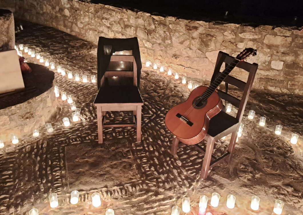 Two wooden chairs sit on a stone courtyard at night, a guitar rests on one chair, candles circle the area.