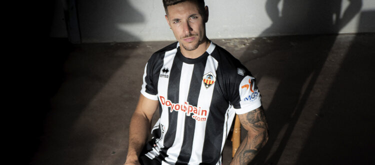 Male soccer player wearing a black-and-white striped jersey sits in a sunlit, shadowed room, looking at the camera with a tattooed arm visible.