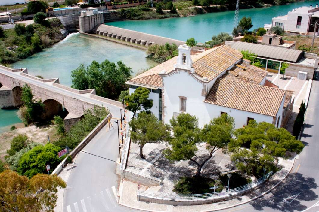White chapel with a tiled roof sits on a small plaza beside a turquoise river and stone bridge (aerial view).