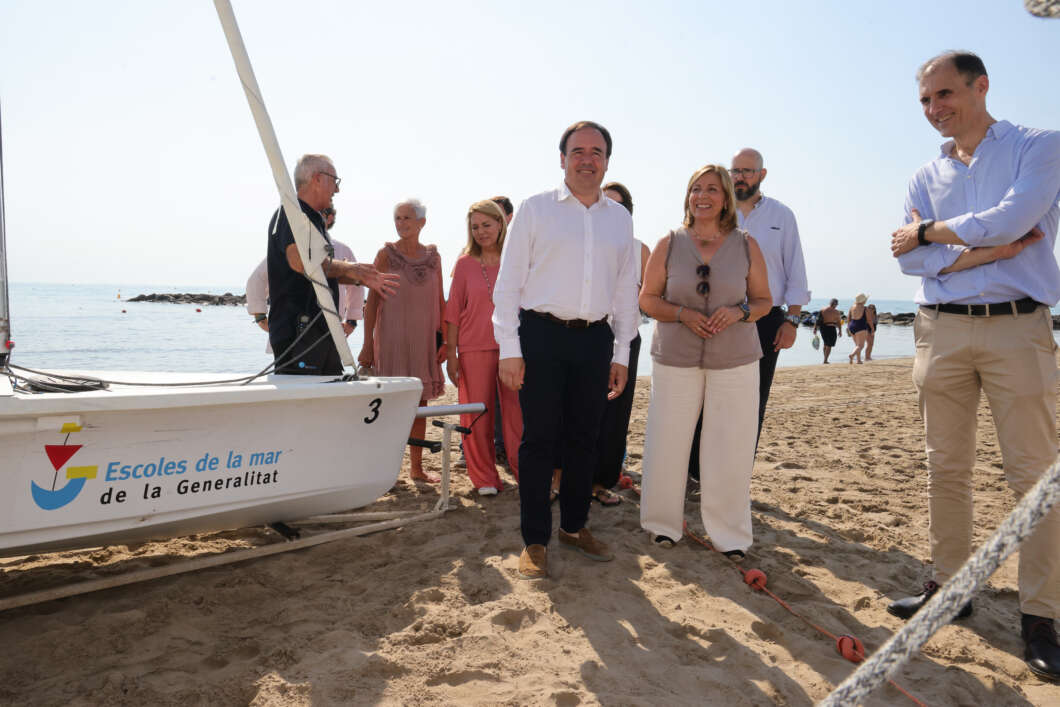 Group of adults standing on a sunny beach beside a small sailboat branded 'Escoles de la mar de la Generalitat'