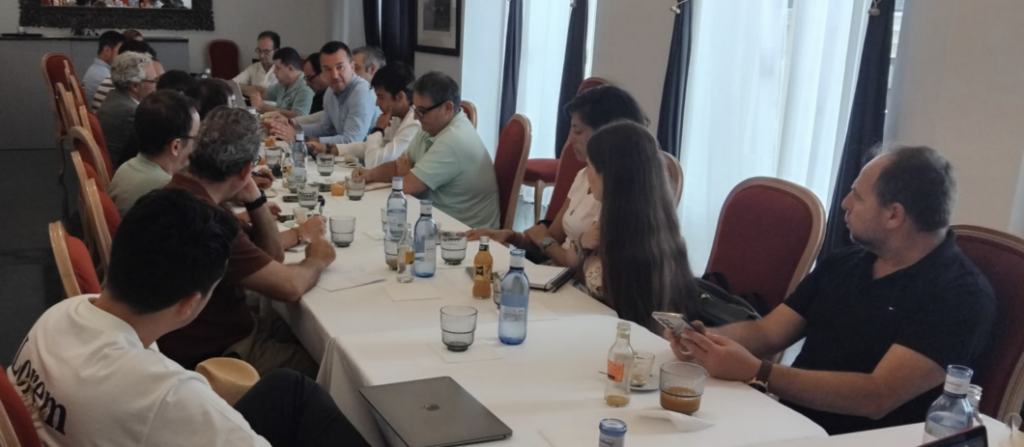 A conference table with many people seated and discussing, bottles of water and papers on the table in a bright meeting room.