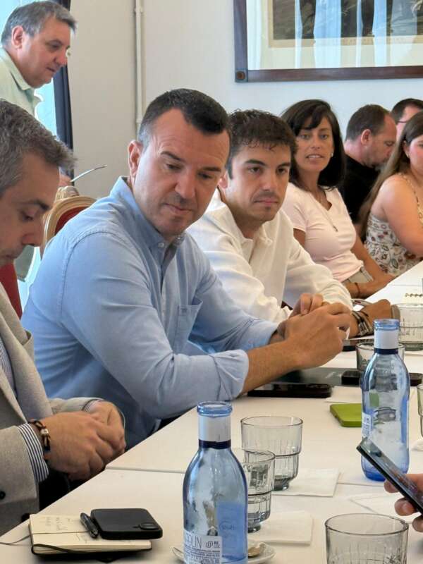 Group of professionals seated around a conference table in a meeting; man in a light blue shirt in the foreground looks down as others listen.