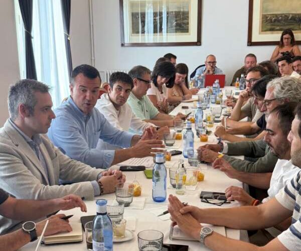 Group of professionals seated around a long conference table, engaged in a discussion, with drinks and notebooks visible on the table.