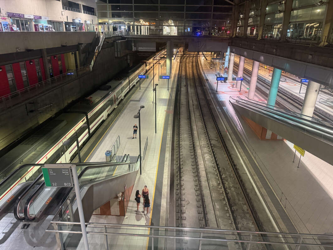 Bird's-eye view of a modern train station at night: a train on the left platform, empty tracks in the center, and a pedestrian bridge with people on the right.