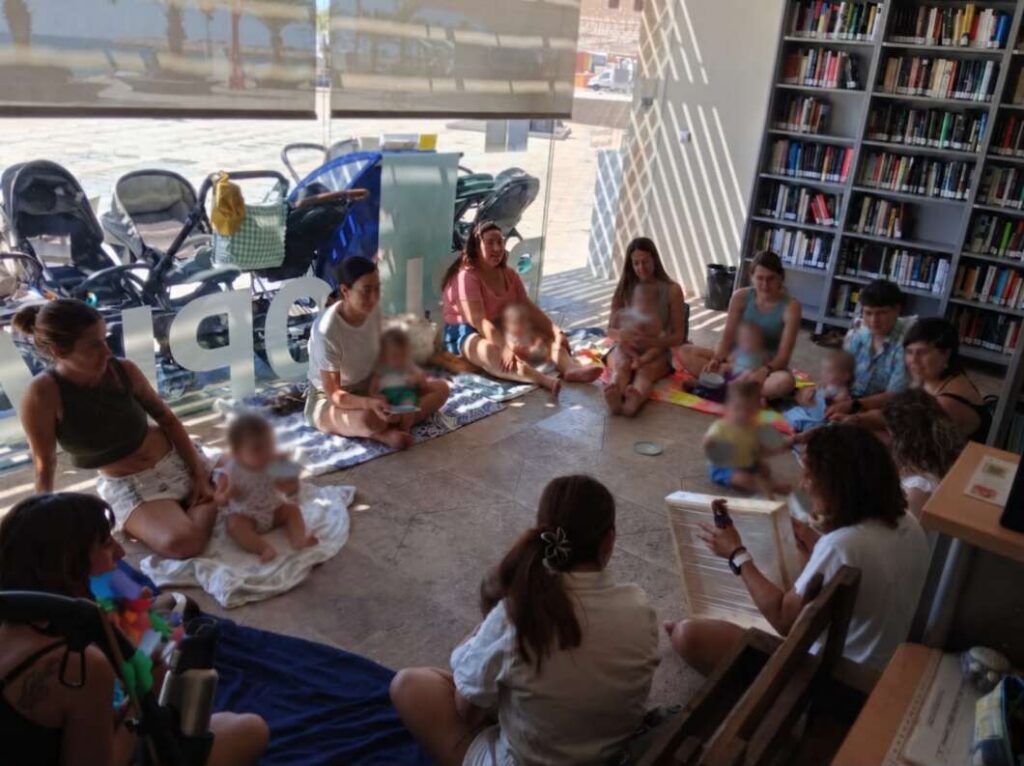 Storytime circle in a bright library: adults and babies sit on blankets in a circle as a reader shows a book on a chair near shelves of books.