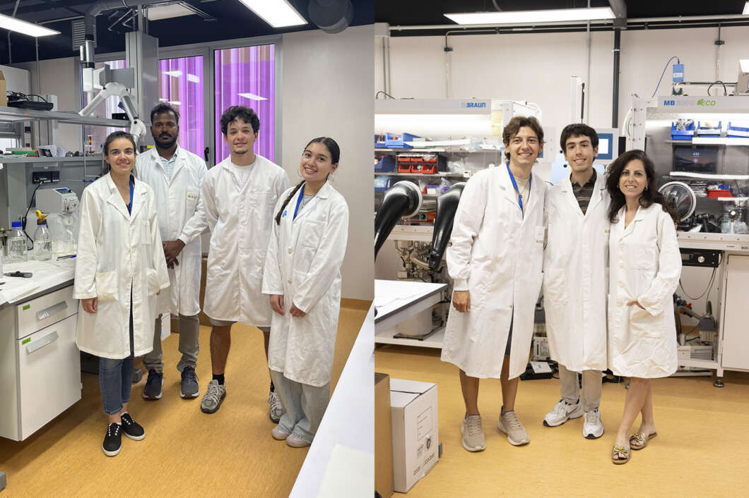 A diverse team of eight scientists in white lab coats posing together in a modern laboratory with workbenches, glassware, and high-tech equipment in the background.