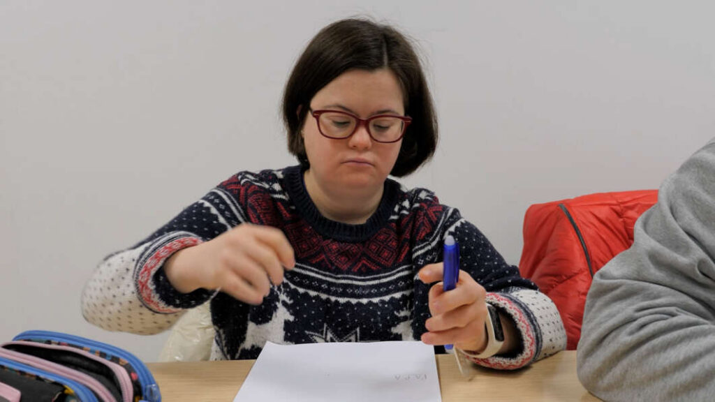 Girl with short dark hair and red glasses writes on a paper at a desk, holding a blue pen.