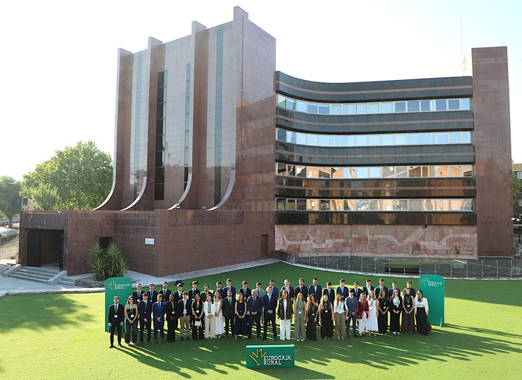 Group of people in formal attire posing for a group photo on a sunny day in front of a modern brick building with green banners visible.