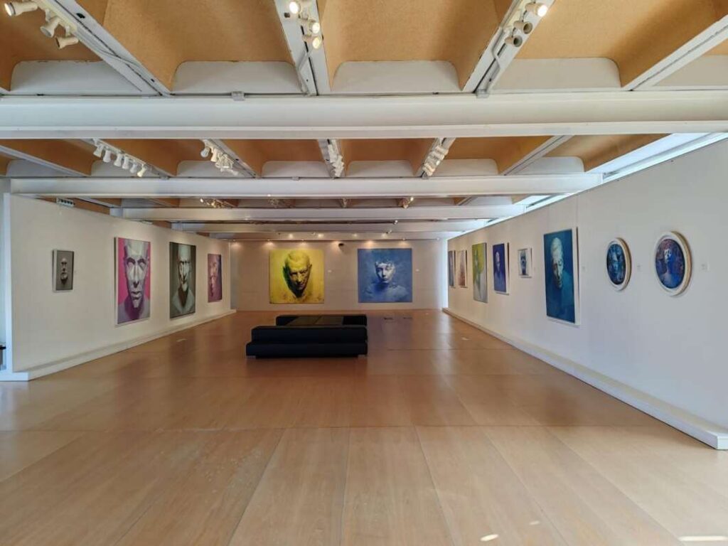 Interior of a bright art gallery with large colorful portrait paintings along white walls and a black bench in the center.