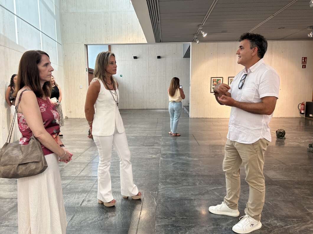 Man in a white shirt and beige pants talks to two women in a modern gallery space with concrete walls.