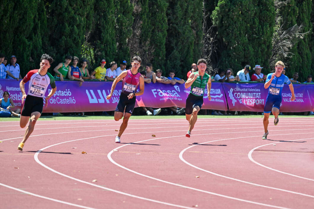 Four male sprinters race on a red track with purple banners and a crowd watching behind the barriers.