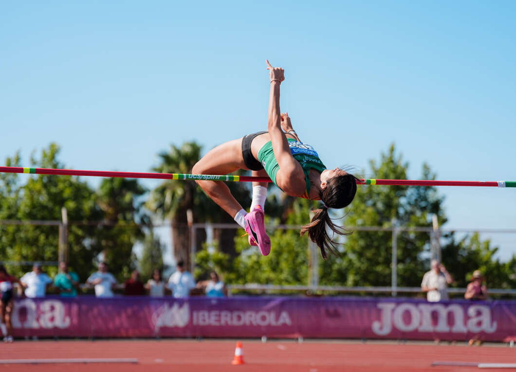 Female high jumper in a green outfit clears a bar during an outdoor track meet on a sunny day; pink shoes and a ponytail visible.
