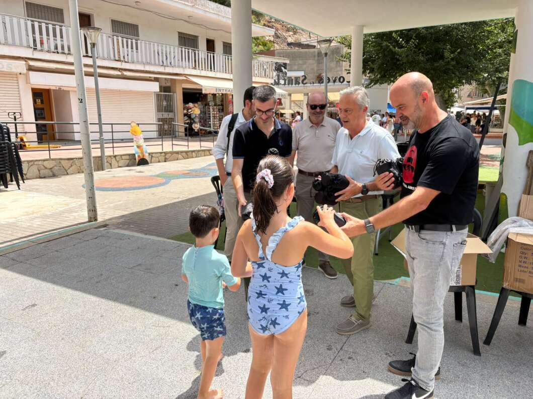 Group of adults giving cameras to two children at an outdoor event, with onlookers nearby.
