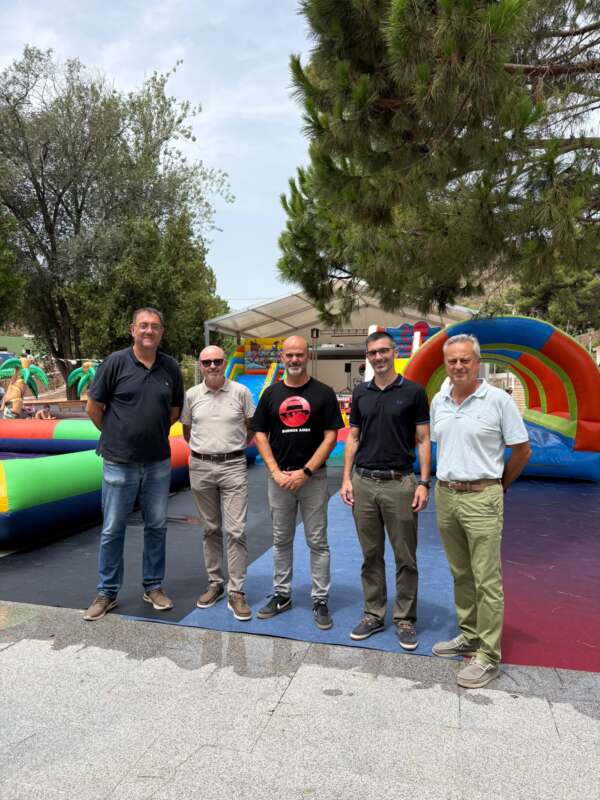 Five men stand together outdoors in front of a colorful inflatable obstacle course at a fair or event.