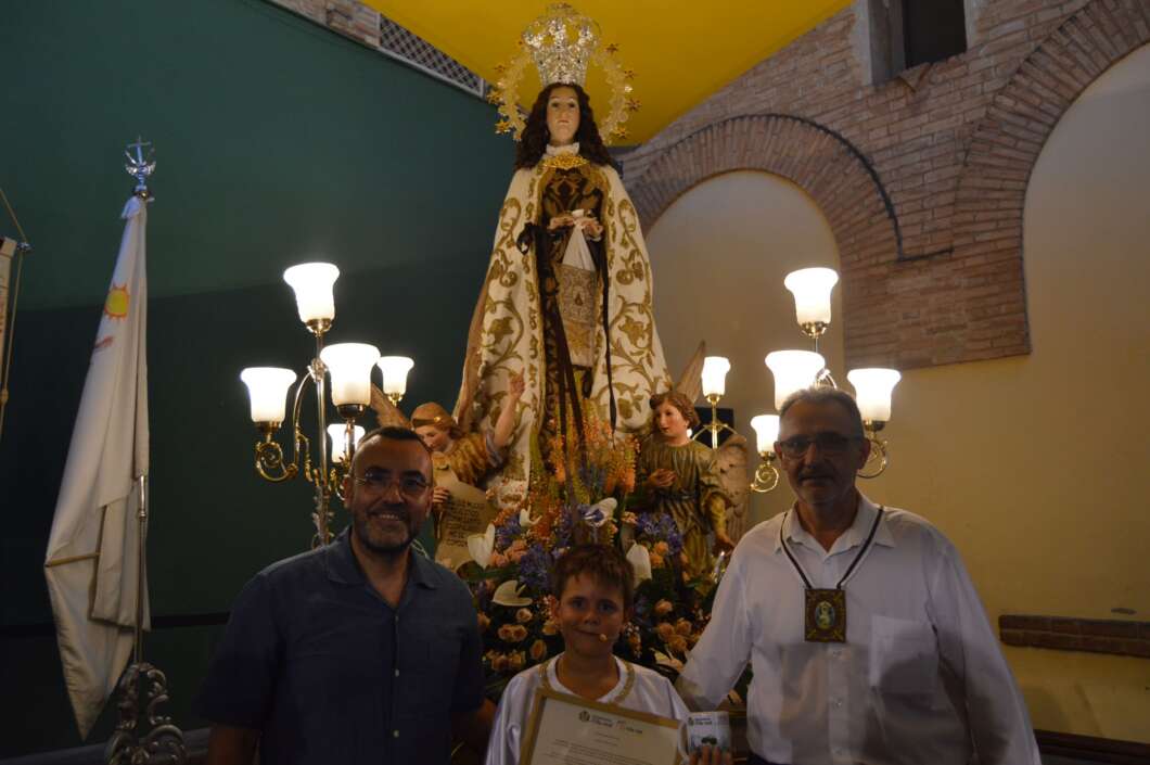 Religious statue of Mary with attendants and two men and a boy holding a certificate in a festively lit hall.