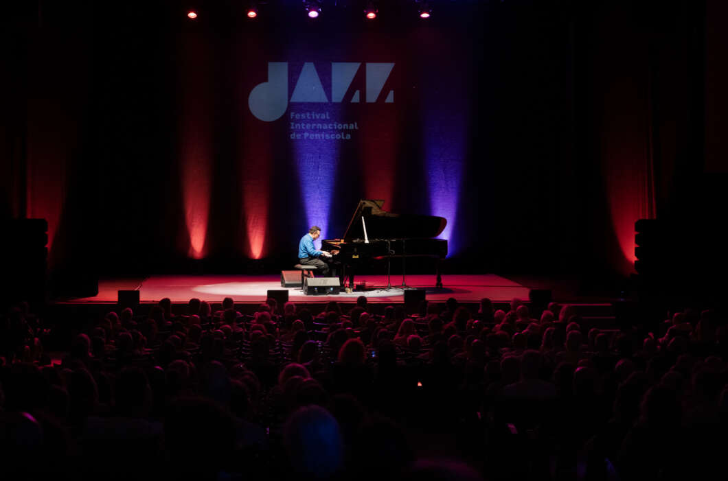 A pianist in a blue shirt performing on a grand piano on a dimly lit stage with a red and blue backdrop and an audience watching the concert.