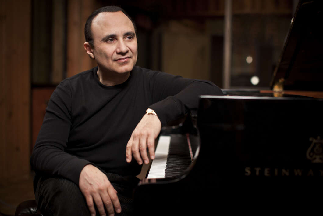 Man in a black long-sleeve shirt seated at a Steinway grand piano in a dimly lit studio. (informative)
