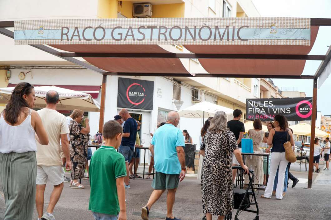 Outdoor food festival scene with people walking under a sign that reads 'RACO GASTRONOMIC' and stalls in the background under a wooden canopy.