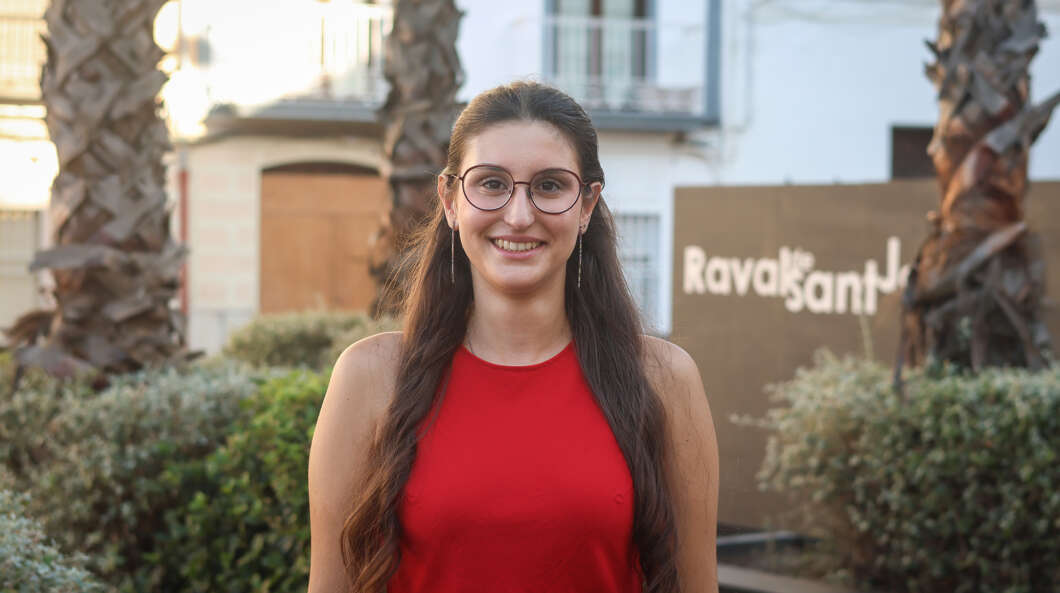 Smiling woman with long dark hair and round glasses, wearing a red sleeveless top, outdoors with greenery and a sign in the background.
