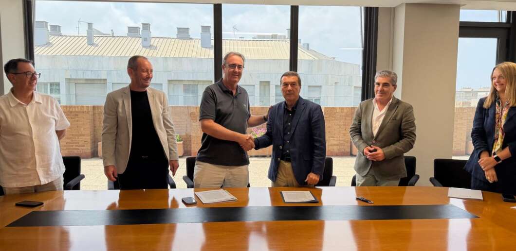 Six professionals stand around a conference table; two men shake hands after signing documents in a bright meeting room, with others watching.