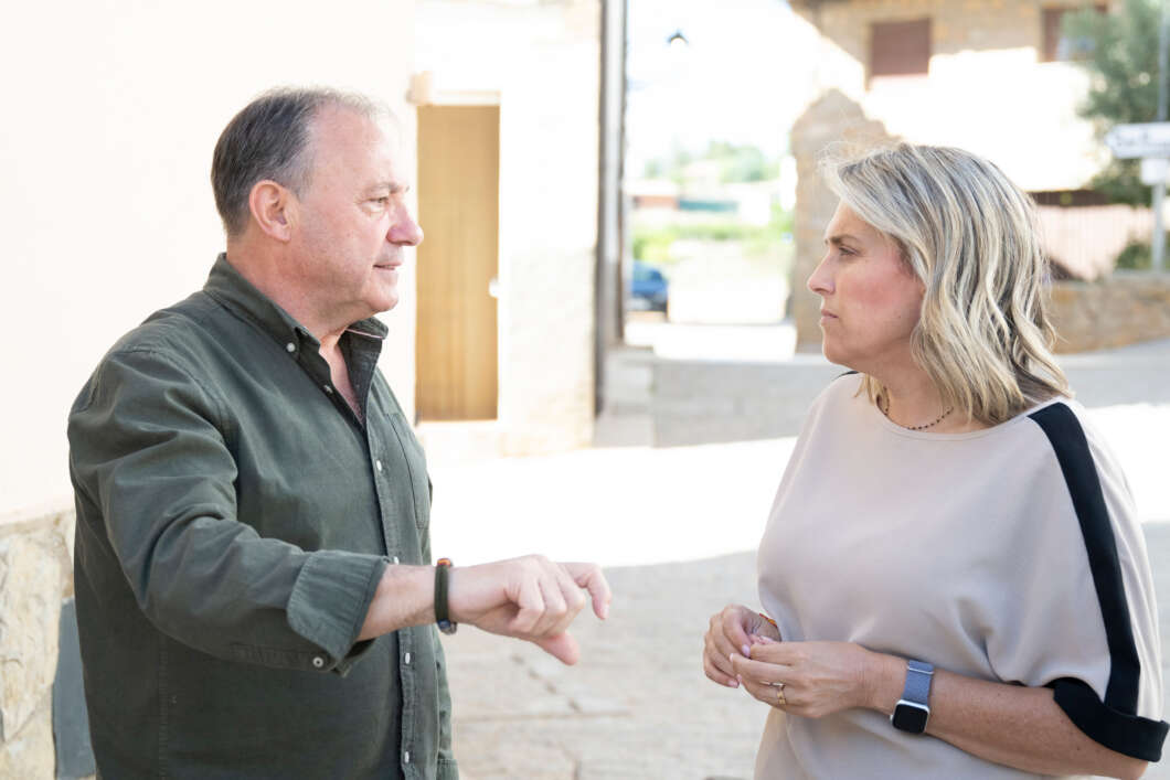 Two adults converse outdoors on a sunny street, the man gesturing with his hand as the woman listens near light-colored buildings.