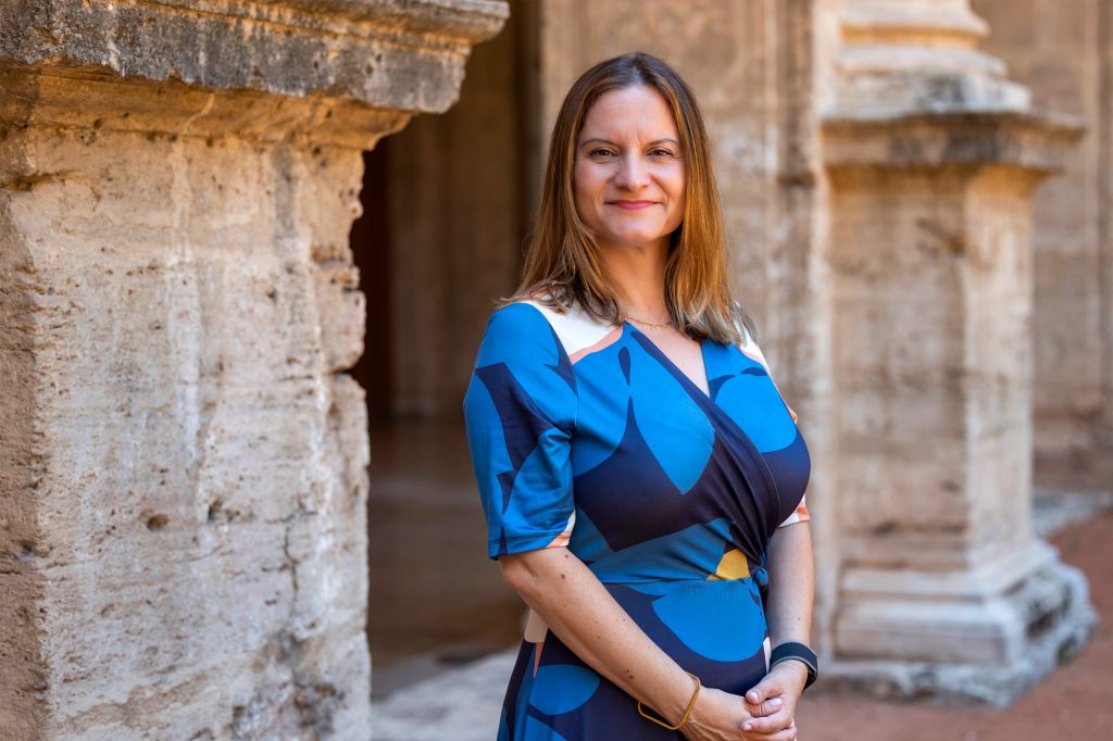 Portrait of a woman in a blue patterned dress standing beside ancient stone columns, smiling at the camera.