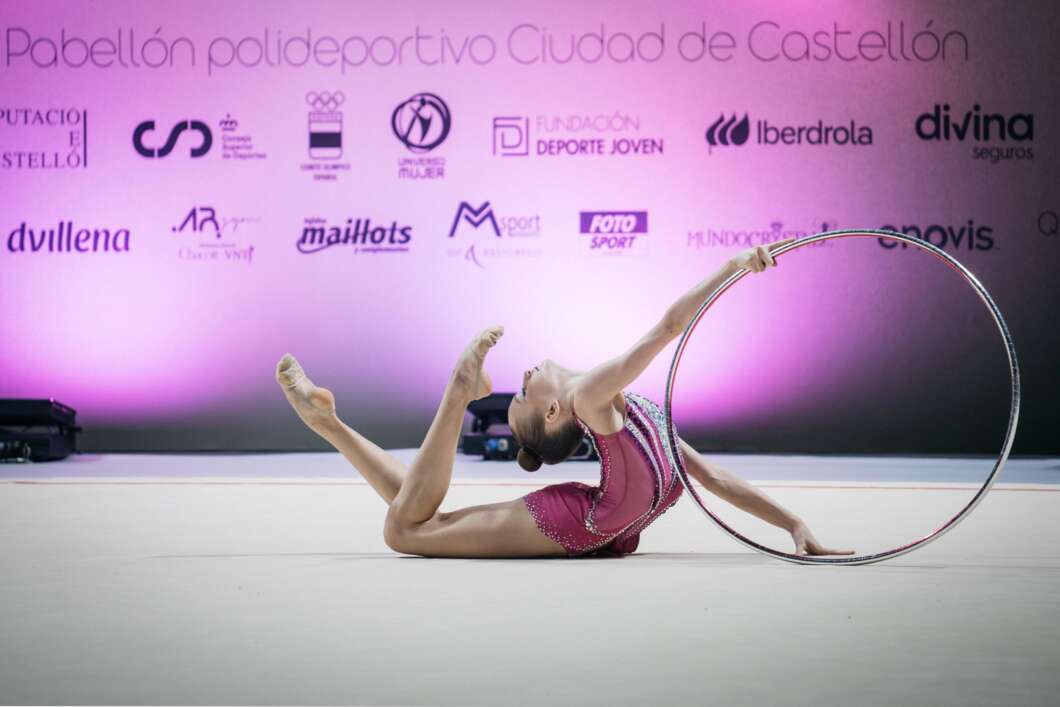 Rhythmic gymnast in a magenta leotard performing a hoop routine on a competition floor with a pink sponsor backdrop.