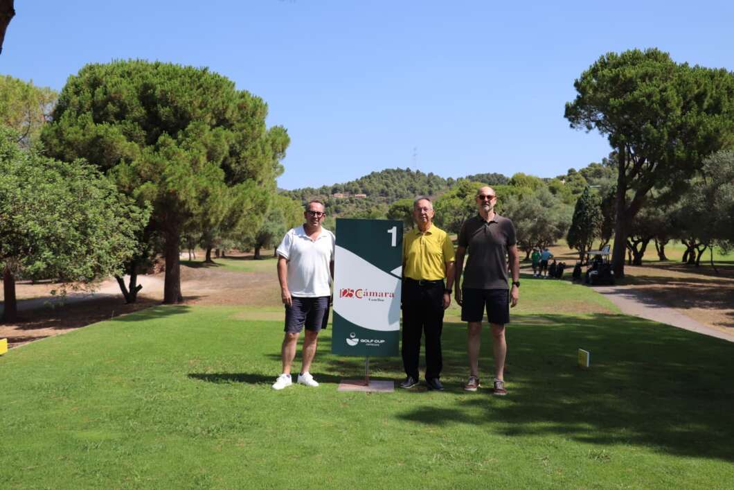 Three men standing on a sunny golf course beside a large numbered sign (1) with logo, trees and a blue sky in the background.