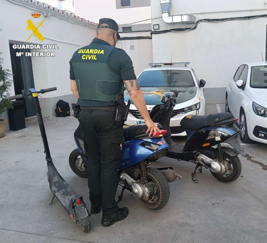 Guardia Civil officer in uniform inspecting two parked motorcycles in a courtyard with a patrol car behind.