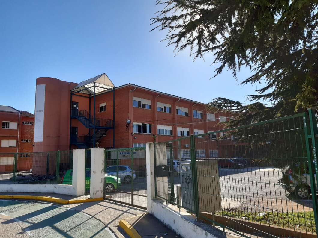 Red brick multi-story building behind a green metal fence with a gated entrance and a blue exterior staircase on sunny day.