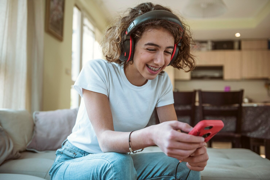 Teen girl with braces wearing black and red over-ear headphones, smiling at a red smartphone while sitting on a sofa at home.