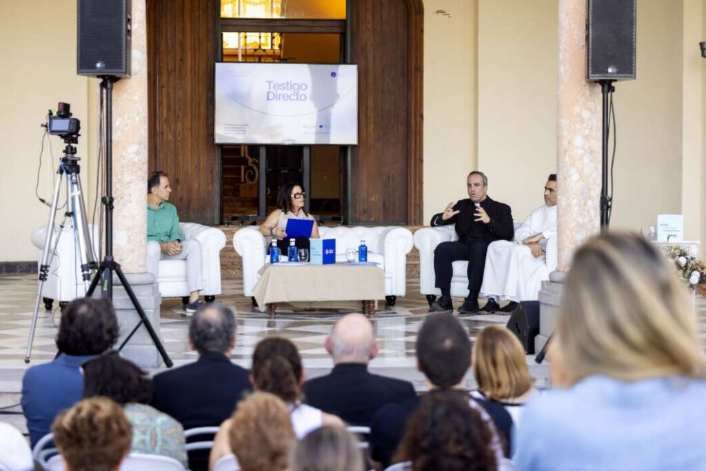 Panel discussion with four speakers seated on white chairs; audience watches as cameras capture the event, backdrop shows Testigo Directo.
