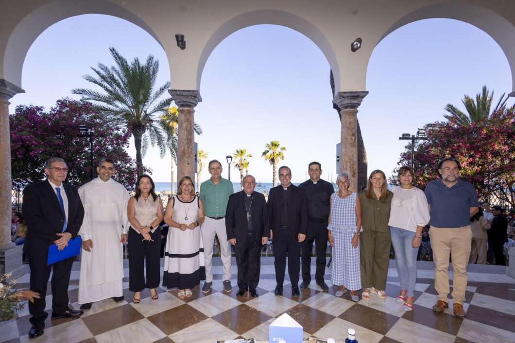 Group of fifteen people, including priests and civilians, posing for a formal photo under arched columns on a seaside terrace with palm trees and flowering bushes.
