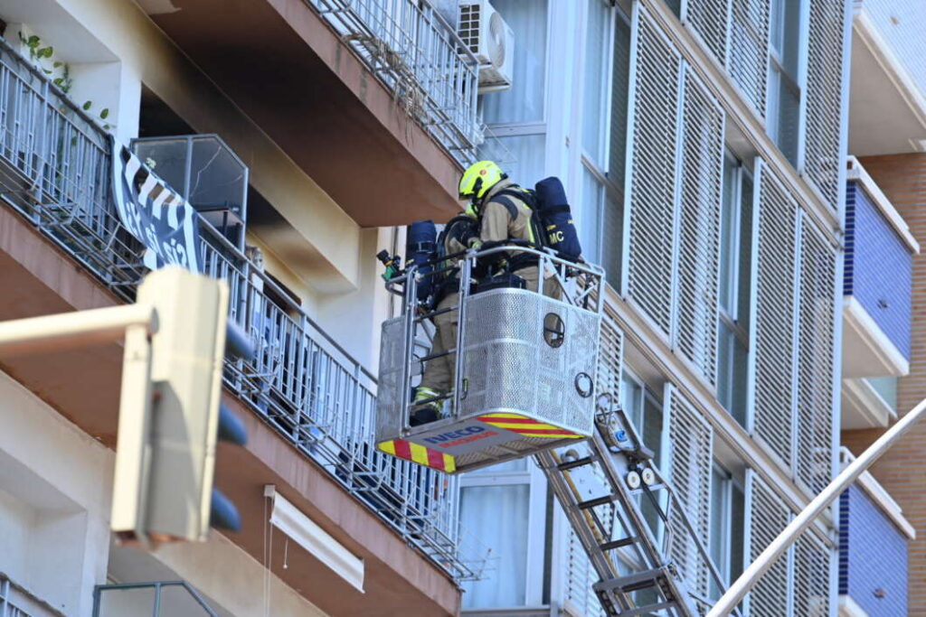 Firefighter in full gear standing in a raised rescue basket outside a multi-story apartment building.