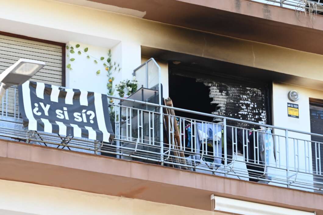 Apartment balcony with a black-and-white striped banner reading '¿Y si sí?' draped over the railing, and clothes hanging on a line beside potted plants and an open door.