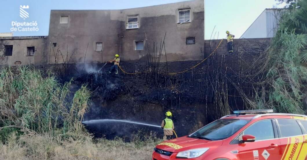 Firefighters extinguishing a brush fire at the base of a concrete building, with a red emergency vehicle nearby.