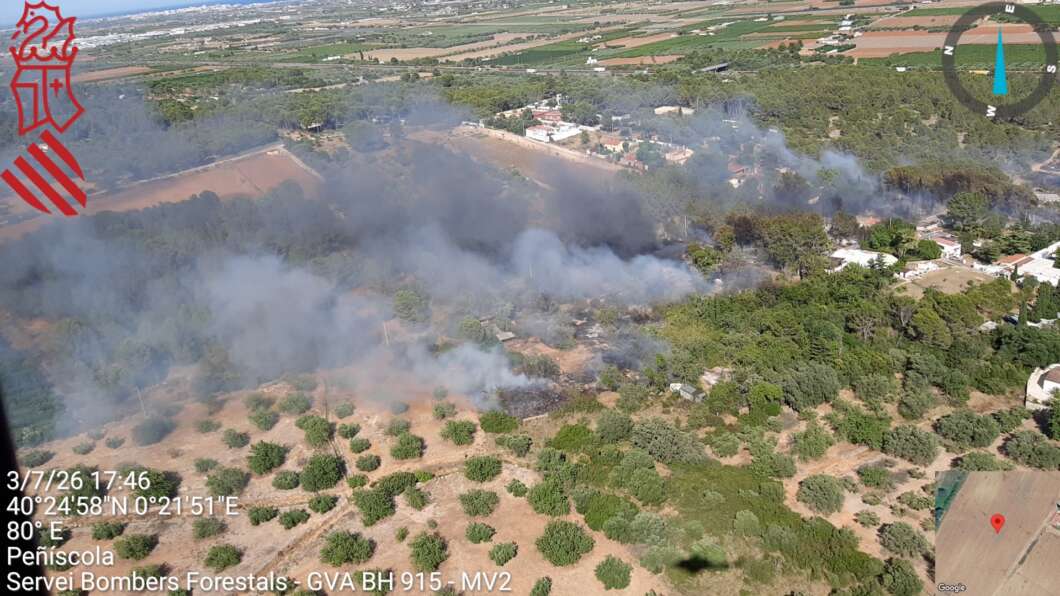 Aerial view of a forest fire with smoke plumes drifting over orchards and a small town; emergency service logo in the corner and coordinates overlayed on screen.