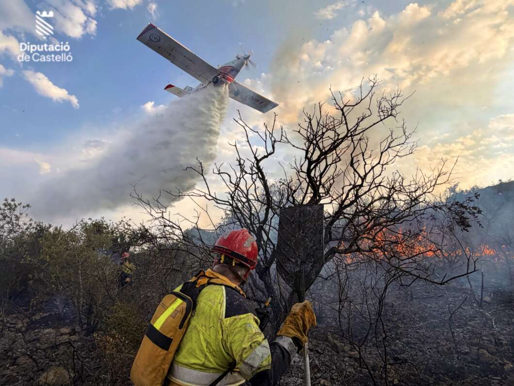 Firefighter in yellow gear and red helmet stands amid a charred hillside as a water-dropping plane streams water onto a wildfire; smoke and flames in the background; Diputació de Castelló logo visible in the sky.