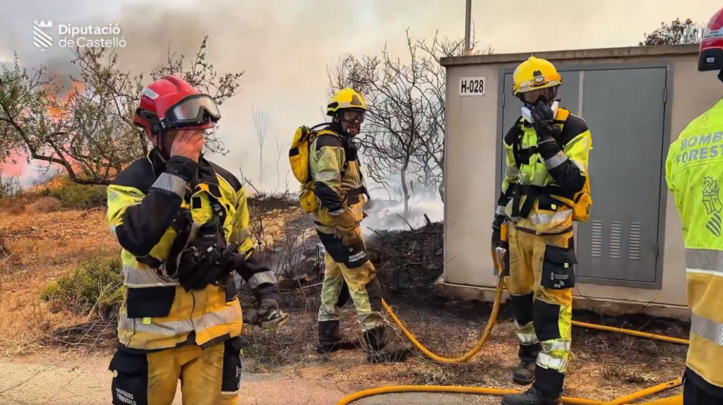 Firefighters in yellow protective gear coordinate a brush fire suppression, holding hoses near a metal cabinet with smoke in the background.