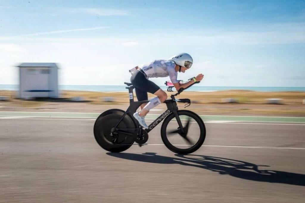 Cyclist in a white jersey and helmet riding a black Cervelo time-trial bike in a low aero position along a coastal road with blurred background.
