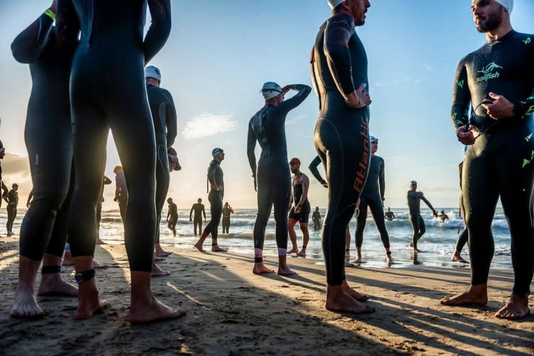 Group of swimmers in wetsuits stand on a sandy beach at dawn, preparing near the ocean.