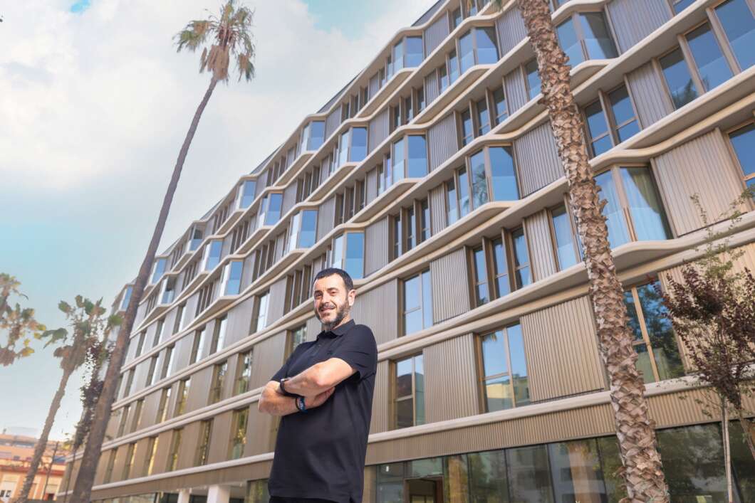 Smiling man with arms crossed in front of a modern glass-and-tile apartment building with palm trees nearby.