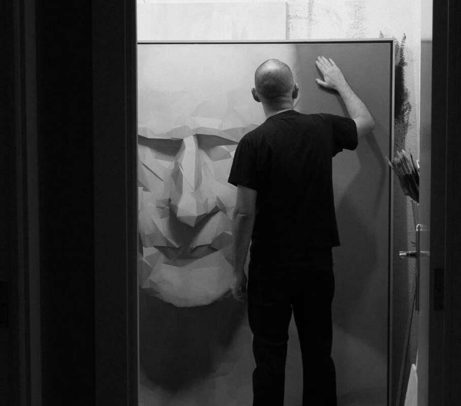 Back view of a person in a black shirt pressing a geometric paper-face sculpture on a wall in a studio workshop.