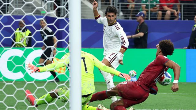 Soccer moment: a player in white shoots as the goalkeeper in neon dives to block near the goal line, a red-clad attacker nearby.