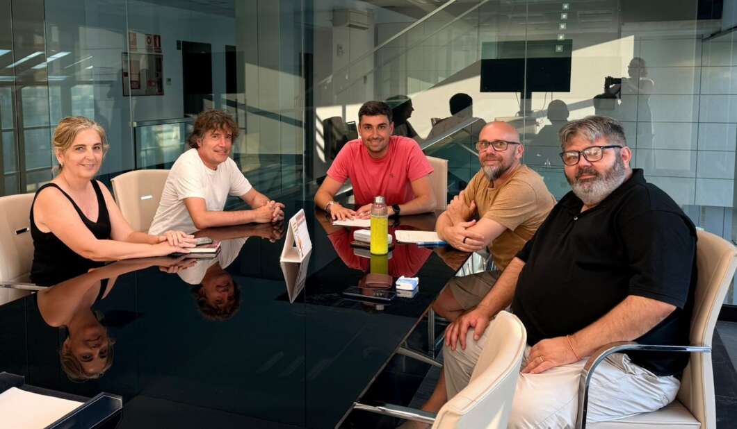 Five people in casual clothes sit around a glass conference table in a modern office, smiling at the camera.