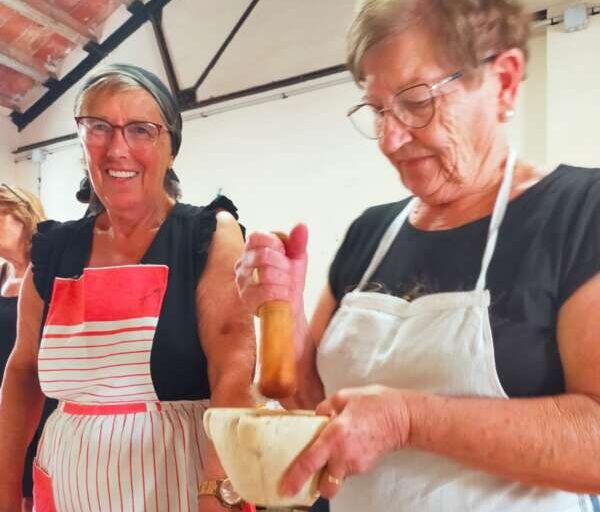 Two older women in aprons in a kitchen, one grinds with a pestle in a mortar while the other smiles and watches.