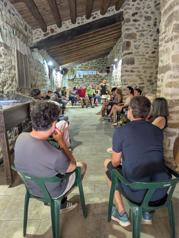 People sit in plastic chairs forming a large circle inside a stone-walled room with wooden beams, engaged in a group discussion.