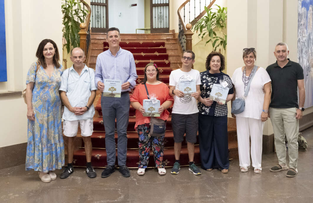 Group of eight adults standing on a red-carpet staircase in a building lobby, some holding booklets or brochures.